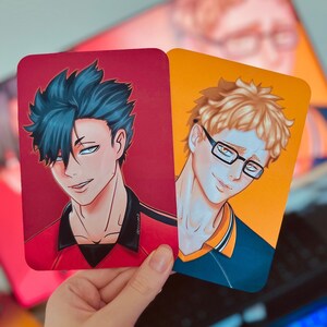 Puede incluir: Dos impresiones art&iacute;sticas rectangulares con ilustraciones de estilo anime de personajes masculinos. Una impresi&oacute;n tiene un fondo rojo y un personaje con cabello azul oscuro. La otra impresi&oacute;n tiene un fondo naranja y un personaje con cabello rubio y gafas.