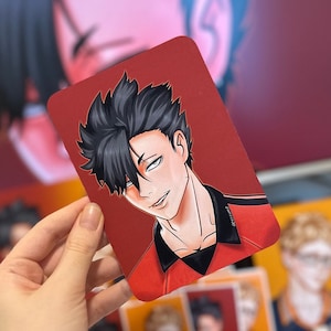 Puede incluir: Una impresi&oacute;n art&iacute;stica rectangular roja que presenta un personaje de anime masculino con cabello negro y un atuendo rojo y negro. El personaje tiene una expresi&oacute;n segura. La impresi&oacute;n es sostenida por una mano, con otras impresiones visibles en el fondo.