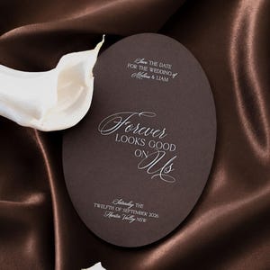 May include: An oval-shaped dark brown wedding save the date card featuring white text that reads "SAVE THE DATE FOR THE WEDDING OF Melina & LIAM", "Forever LOOKS GOOD ON Us", and "Saturday THE TWELFTH OF SEPTEMBER 2026 Hunter Valley NSW" next to a white calla lily.