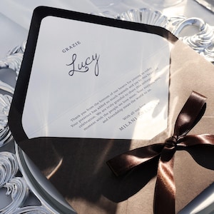 May include: A wedding invitation with a white card featuring the name "Lucy" in cursive and the word "GRAZIE" at the top. The card is inside a brown envelope tied with a brown satin ribbon. The invitation is on a decorative silver plate.