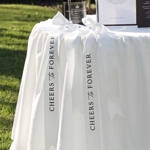 Puede incluir: Un mantel redondo blanco con dos cintas largas de satén blanco que cuelgan por el borde. Cada cinta está atada con un lazo en la parte superior y tiene impreso el texto negro "CHEERS TO FOREVER" en orientación vertical a lo largo.