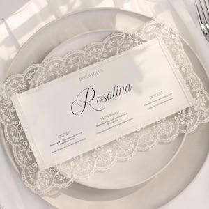 Może przedstawiać: Biała karta menu z napisem "Rosalina" w eleganckiej czcionce, obramowana delikatną koronkową bordiurą, spoczywa na białym talerzu. Srebrne sztućce i biały obrus dopełniają elegancką scenę.