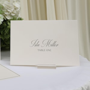 Peut inclure: Carton de table ivoire avec le nom "Isla Miller" et "Table One" imprimés en script noir élégant. La carte est présentée dans un support en acrylique transparent, avec un arrangement floral en arrière-plan. Papeterie de mariage ou d'événement.
