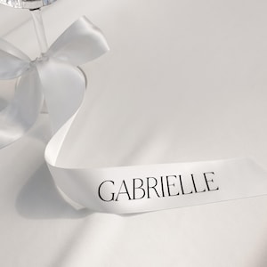 May include: A white satin ribbon with the name "GABRIELLE" printed in black script. The ribbon is draped across a white surface, with a glass stem in the background. A white bow is tied around the stem.