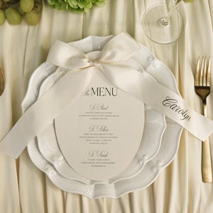 Könnte beinhalten: Eine formelle Tischdekoration mit einem weißen Band, das um einen Stapel weißer Teller gebunden ist. Eine Menükarte ist unter dem Band versteckt. Die Menükarte lautet "The Menu" und listet die Gänge auf. Eine goldene Gabel, ein Messer und ein Löffel sind sichtbar.