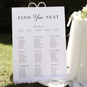May include: A white wedding seating chart with black text. The chart is titled "Find Your Seat" and lists guest names under the numbers one through nine. The chart is displayed on a white easel.