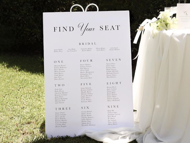 May include: A white wedding seating chart with black text. The chart is titled "Find Your Seat" and lists guest names under the numbers one through nine. The chart is displayed on a white easel.