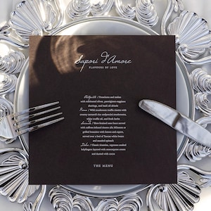 May include: A formal table setting featuring a dark brown menu card with the text "Sapori D'Amore, Flavours of Love" and a detailed menu. The card is placed on a white plate with a decorative silver rim, accompanied by a fork and knife.
