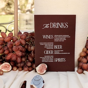 Può includere: Un menu bordeaux con il testo "The Drinks" elenca vini, birra, sidro e liquori. Uva e fichi sono disposti su una tovaglia bianca. Sono visibili anche un bicchiere e un cucchiaio decorativo.