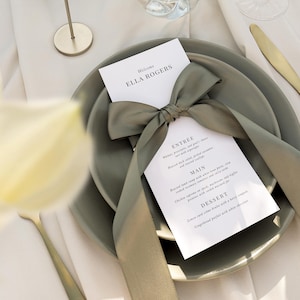 May include: A white card with a green ribbon tied in a bow. The card has the text "Welcome Ella Rogers" and a menu with the headings "Entree", "Main", and "Dessert". The card is on top of two green plates.
