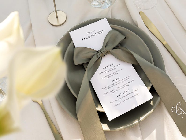 May include: A white card with a green ribbon tied in a bow. The card has the text "Welcome Ella Rogers" and a menu with the headings "Entree", "Main", and "Dessert". The card is on top of two green plates.