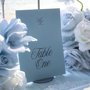 Könnte beinhalten: Hellblaue Tischkarte mit dem Schriftzug "Table One" in eleganter Schrift, verziert mit den Initialen "AL". Die Karte ist von hellblauen und weißen Rosen umgeben, wodurch eine weiche, romantische Atmosphäre entsteht, ideal für eine Hochzeit.