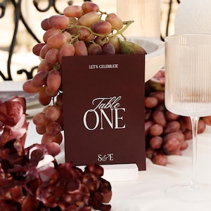 May include: A burgundy table card with the words "Table ONE" in white script, and the text "LET'S CELEBRATE" above. The card is propped up by a white block, with grapes and a clear glass nearby. The scene is set on a white tablecloth.