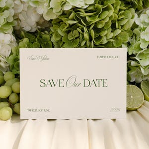 Könnte beinhalten: Eine cremefarbene "Save Our Date" Karte mit eleganter grüner Schrift, umgeben von grünen und weißen Hortensien, Trauben und Limettenscheiben. Die Karte enthält die Namen "Emie & Julian", das Datum "Twelfth of June 2026" und den Ort "Hawthorn, VIC".