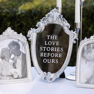 May include: Silver framed decor with the text "THE LOVE STORIES BEFORE OURS". Two framed photos of an elderly couple are on either side of the sign. Two white candles are in the background.