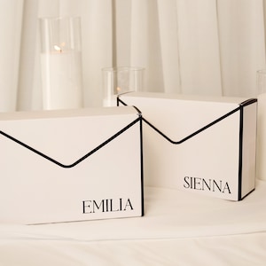 Pode incluir: Duas caixas de presente retangulares brancas em forma de envelope com bordas pretas. A caixa à esquerda apresenta o nome EMILIA impresso em preto, e a caixa à direita apresenta o nome SIENNA impresso em preto no canto inferior direito.
