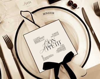 Peut inclure: Une table dressée avec une nappe blanche et une carte de menu noir et blanc qui dit "Bon Appetit" avec un ruban noir autour. La carte de menu liste "Canapes", "Starter", "Main", et "Dessert". La table est également dressée avec une serviette blanche, une assiette noire, une fourchette, un couteau et un verre.