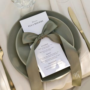 May include: A white card with the text "Welcome Ella Rogers" and a menu for an event. The card is tied with a sage green ribbon and placed on top of two sage green plates. A gold fork and knife are on the table.