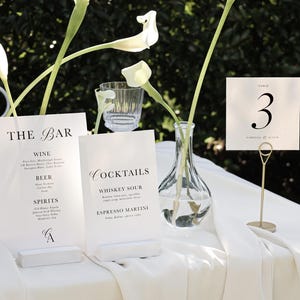 May include: White table with a white tablecloth, two white signs with black text, a clear glass vase with white flowers, and a gold table number holder with a white card that says "Table 3". The signs list the bar menu with drinks like wine, beer, spirits, whiskey sour, and espresso martini.