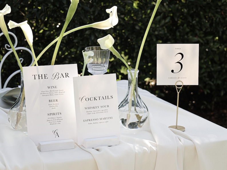 May include: White table with a white tablecloth, two white signs with black text, a clear glass vase with white flowers, and a gold table number holder with a white card that says "Table 3". The signs list the bar menu with drinks like wine, beer, spirits, whiskey sour, and espresso martini.