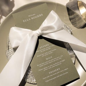 May include: A green card with white text on it, tied with a white satin ribbon. The text reads "Welcome Ella Rogers" and lists a menu with the headings "Entree", "Main", and "Dessert".