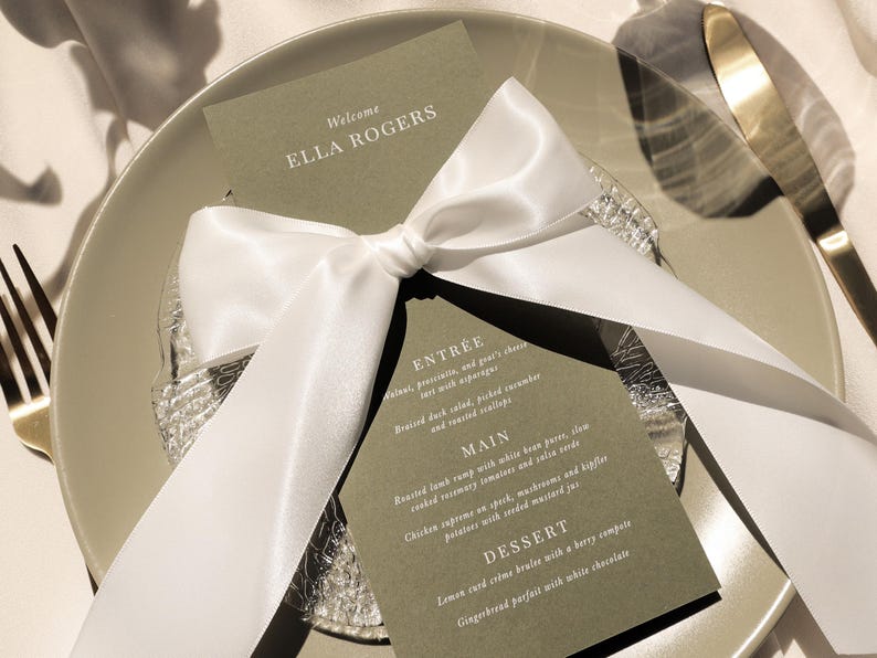May include: A green card with white text on it, tied with a white satin ribbon. The text reads "Welcome Ella Rogers" and lists a menu with the headings "Entree", "Main", and "Dessert".