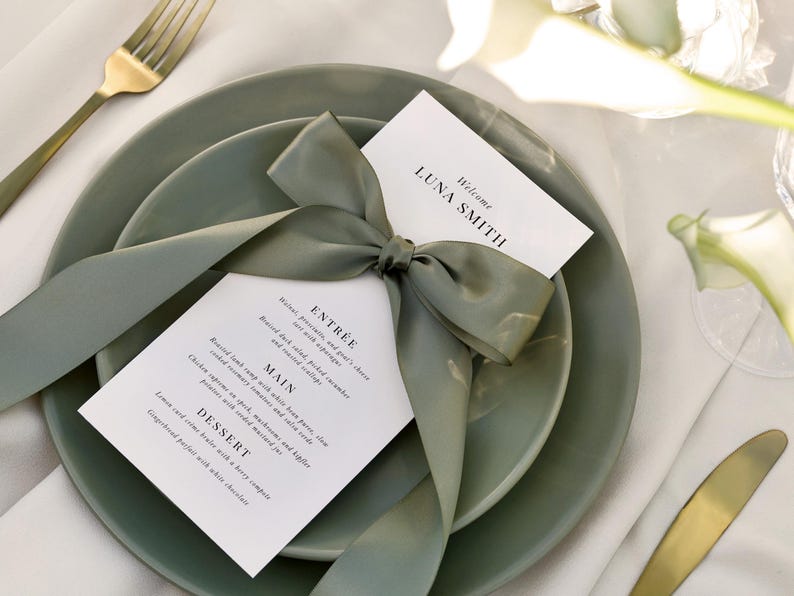 May include: A white card with a green ribbon tied in a bow. The card has the text "Welcome Luna Smith" and a menu with the headings "Entree", "Main", and "Dessert". The card is on top of two green plates.