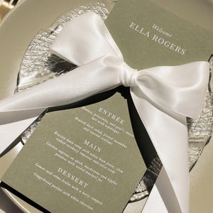 May include: A green card with white text, tied with a white satin ribbon. The card reads "Welcome Ella Rogers" and lists a menu for an event: Entree, Main, Dessert.