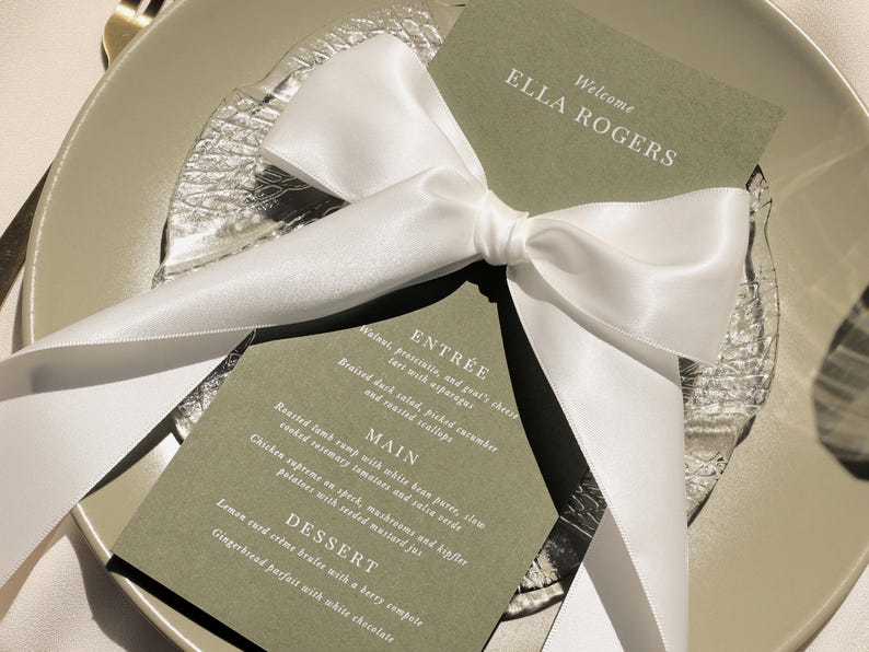 May include: A green card with white text, tied with a white satin ribbon. The card reads "Welcome Ella Rogers" and lists a menu for an event: Entree, Main, Dessert.