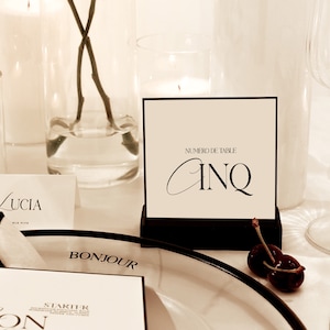 Puede incluir: Una tarjeta de número de mesa en blanco y negro con el texto "NUMERO DE TABLE" y "CINQ" en un soporte negro. La tarjeta está sobre una mesa blanca con una servilleta blanca, un vaso de agua y una vela encendida.
