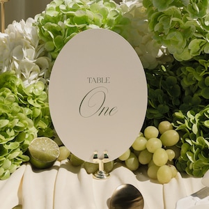 以下が含まれることがあります： エレガントな筆記体で「TABLE One」と書かれたアイボリー色の楕円形のテーブルナンバーカード。カードは金のスタンドに立てられ、緑と白の花、ブドウ、ライムの半分に囲まれています。クリーム色のテーブルクロスの上にシーンが設定されています。