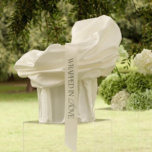 May include: A white, draped fabric sculpture with the words "Wrapped in Love" on a ribbon. The sculpture sits on a clear pedestal, with a bouquet of white and green flowers nearby. The background is a blurred green landscape.
