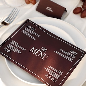 Może przedstawiać: Nakrycie stołu z białym talerzem, sztućcami i kartą menu. Karta menu jest ciemnobrązowa z białym tekstem, szczegółowo opisującym menu. Obecna jest również karta z imieniem "Olivia".