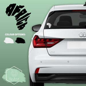 May include: A white car with a black "Africa" decal on the back window. The decal is shaped like the continent of Africa and the word "Africa" is spelled out within the outline of the continent. The car is parked on a green background.
