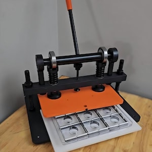 May include: A manual die cutting machine featuring a black metal frame, an orange pressure plate, and a vertical orange handle. The device includes two vertical springs, a horizontal bar with ball bearings, and a clear plastic grid mould with six circular cutouts.