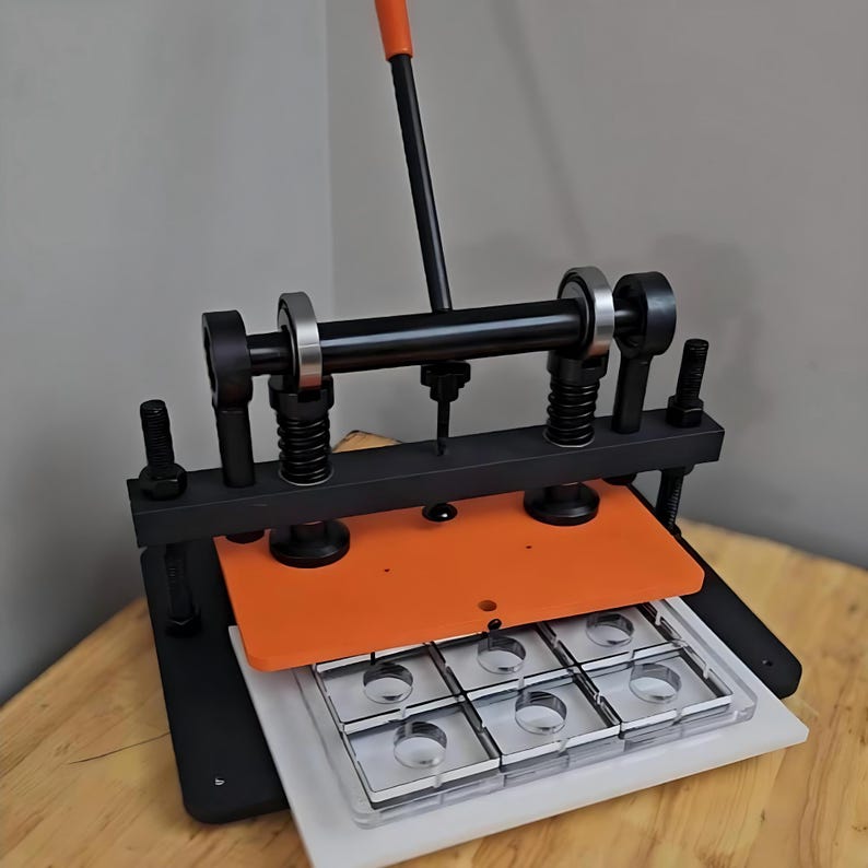 May include: A manual die cutting machine featuring a black metal frame, an orange pressure plate, and a vertical orange handle. The device includes two vertical springs, a horizontal bar with ball bearings, and a clear plastic grid mould with six circular cutouts.