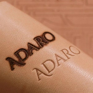 May include: Close-up of a light brown leather surface with the word "ADARO" embossed twice, once in a larger, more stylized font and once in a smaller, more traditional font.