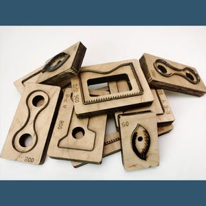 May include: A collection of light-coloured wooden die-cutting blocks featuring various metal blade shapes embedded in the surface. Each block is marked with numerical identifiers such as 002, 800, 60, and 200, used for precision cutting of materials.