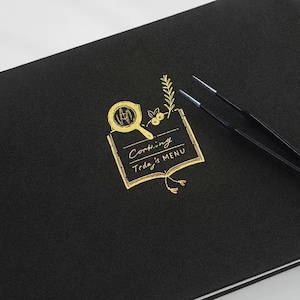 May include: A black notebook with a gold foil design of a frying pan, a sprig of rosemary, and a book with the text "Cooking Today's MENU".