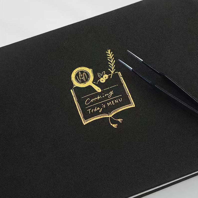 May include: A black notebook with a gold foil design of a frying pan, a sprig of rosemary, and a book with the text "Cooking Today's MENU".