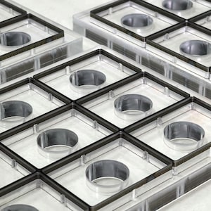 Puede incluir: Primer plano de múltiples moldes de plástico transparente con marcos cuadrados y aberturas circulares. Cada molde tiene un borde gris oscuro. Los moldes están apilados, creando un patrón similar a una cuadrícula. La estética general es limpia e industrial.