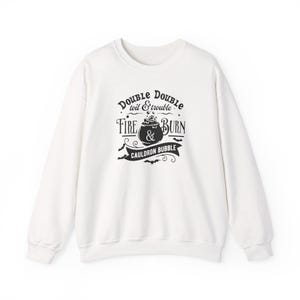 May include: A white long-sleeve sweatshirt featuring a black graphic print on the chest. The design includes a central cauldron illustration surrounded by the text Double Double toil & trouble Fire & Burn Cauldron Bubble and small bats.