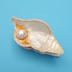 Könnte beinhalten: Eine muschelförmige Haarspange mit perlmuttweißer Oberfläche, goldfarbenen Rändern und einer einzelnen runden, weißen Kunstperle in der Öffnung. Das Accessoire hat eine glatte, glänzende Oberfläche und ein detailliertes, spiralförmiges Design.