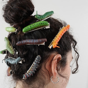 Op de afbeelding: Een persoon met krullend bruin haar draagt ​​verschillende kleurrijke rups haarclips. De clips zijn in verschillende tinten groen, oranje, blauw en rood.