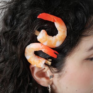 Könnte beinhalten: Zwei Haarspangen mit realistischen Garnelenmotiven. Jede Garnele ist gebogen und weist einen Farbverlauf von Weiß zu Orange-Rosa auf. Sie sind an einem einfachen, goldfarbenen Metallclip befestigt, der im dunklen, lockigen Haar sitzt.