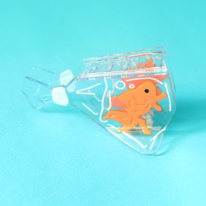 Op de afbeelding: Een doorzichtige plastic paperclip in de vorm van een goudviskom met een oranje goudvis erin. De paperclip is ontworpen om papier bij elkaar te houden.