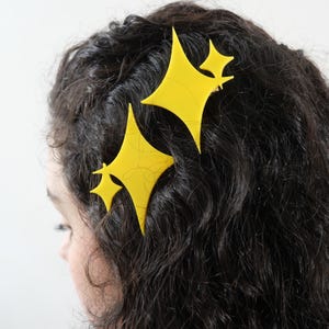 May include: Two bright yellow hair clips shaped like four-pointed stars with smaller star accents attached to the side. The clips are secured in dark, wavy hair, featuring a smooth, matte finish and a distinct geometric design with sharp points.