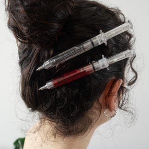 May include: Two hair clips shaped like medical syringes. One clip is clear with black measurement markings, and the other clip contains a red liquid with black measurement markings. Both clips feature a silver-toned tip and a plunger mechanism.