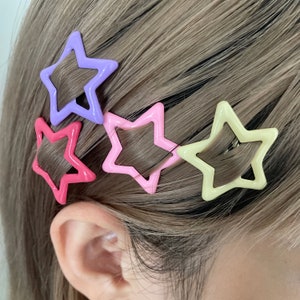 以下が含まれることがあります： 異なる色の星型のヘアクリップが4つあります。紫、ピンク、薄いピンク、黄色です。クリップは人の髪に付いています。