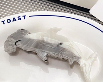 May include: A hammerhead shark-shaped hair claw clip featuring a marbled grey and white finish. The shark design includes a wide head, dorsal fin, and tail, with a smooth, glossy texture. The clip is positioned on a white plate with the word TOAST.